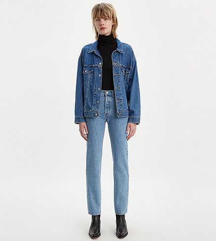 A single model stands in the centre of the image and has a light blonde mullet style haircut and wears a mid wash blue denim trucker jacket over a black turtleneck top that is tucked into jeans. She wears light wash blue 501 Original jeans over a pair of leather black boots.